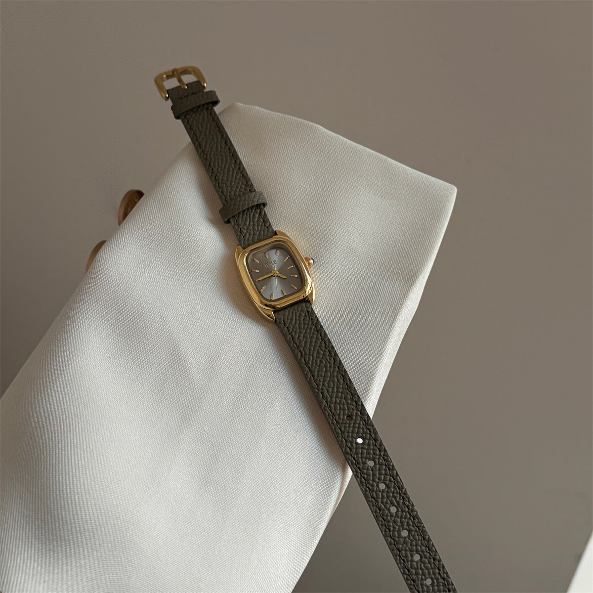 Olive-colored strap watch with a small gold square dial, featuring a niche design